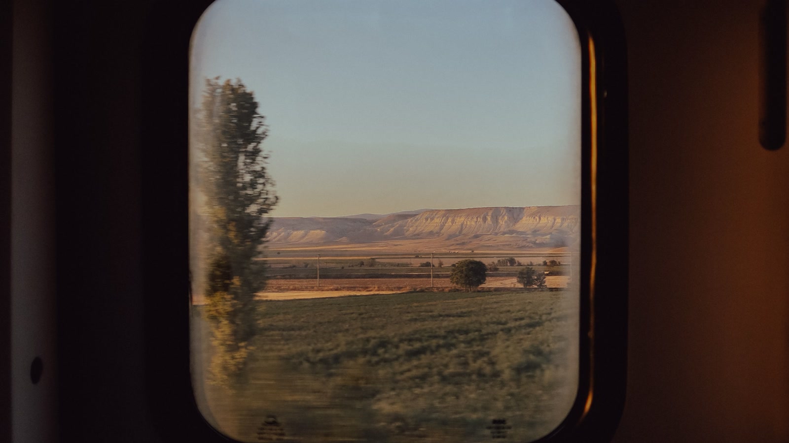 Open arid plains and pale ridges seen through a passenger train window at golden hour.