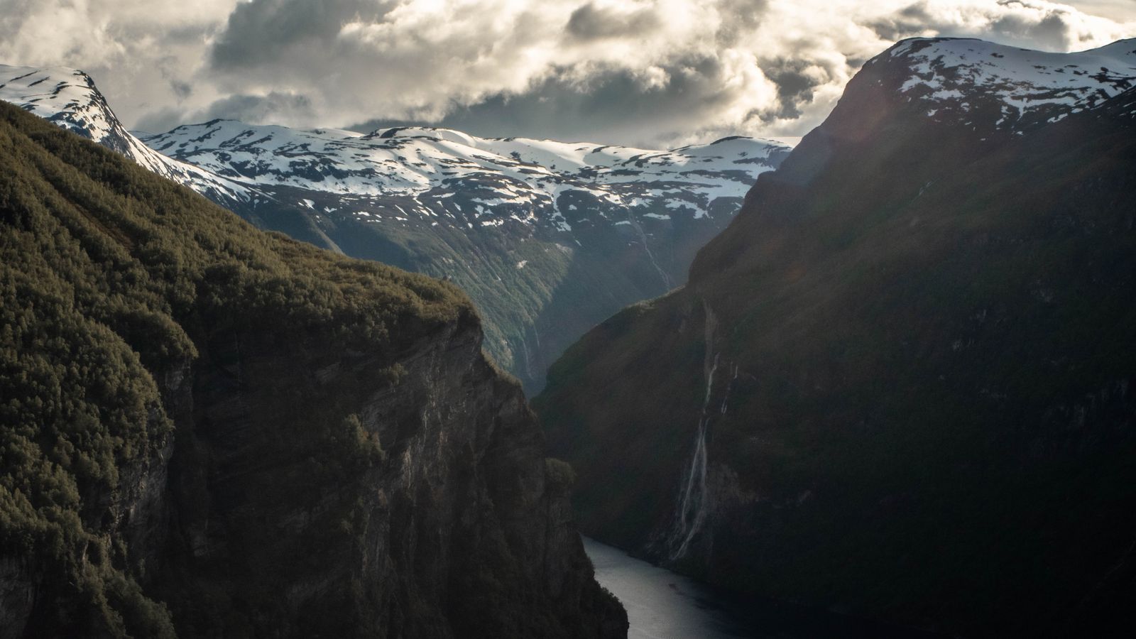 A narrow fjord in Norway between steep cliffs, with snowy peaks and a waterfall.