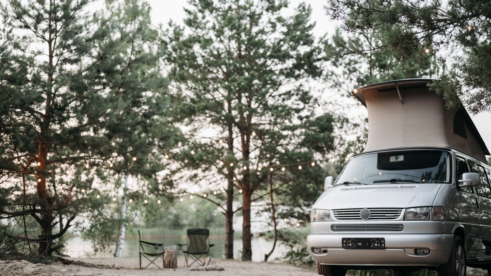A camper van with pop-top roof parked among pines beside water at dusk.