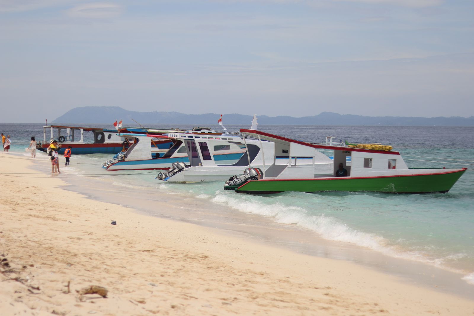 Colorful passenger speedboats and a wooden boat moored in turquoise shallows off a sandy Indonesian beach, with hills on the horizon